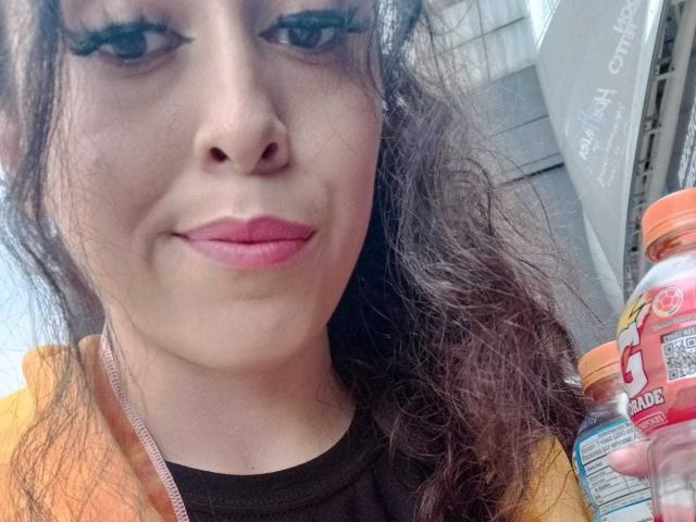 Kendra Simons, com cabelo castanho encaracolado, batom rosa e cílios grossos, segurando uma garrafa de Crystal Light, vestindo uma jaqueta amarela-alaranjada com zíper e uma camisa marrom..