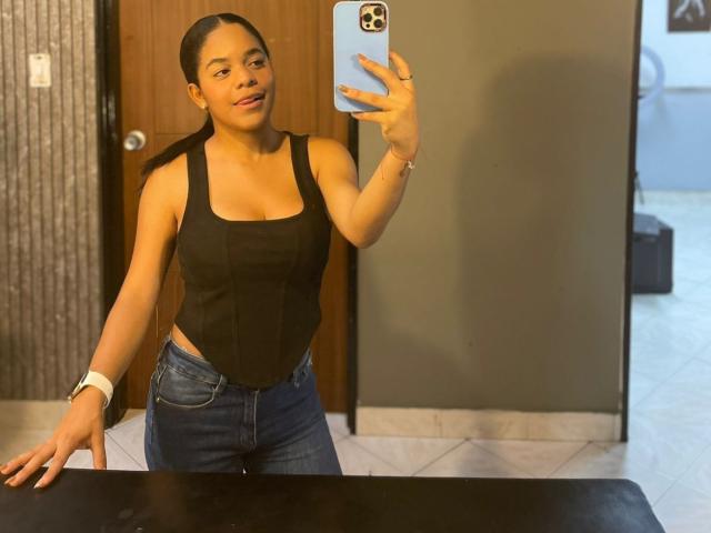 TiffanyyJoness with brown skin, black tank top showing her small tits, blue jeans, and ponytail, taking a mirror selfie, holding a blue phone, wearing a white wristband.