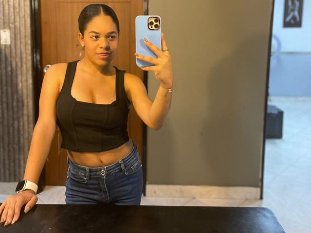TiffanyyJoness with medium brown skin, black square-strap crop top, showing tits, blue jeans, black wristwatch, holding phone, taking selfie in mirror.