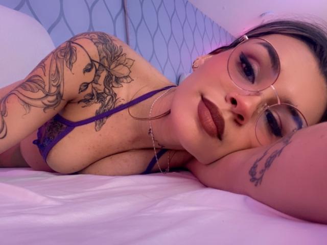 LenaBranch está acostada en una cama, vistiendo lencería púrpura, con gafas, tatuajes en el brazo y el pecho, y lápiz labial oscuro. Sus senos son visibles. Iluminación rosa suave..
