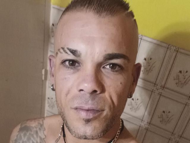 Fonttaka's face with a shaved mohawk, dark eyebrows, small nose, tattoos on the shoulder, chain necklace, and earrings, against a yellow wall. His expression is neutral and slightly serious.