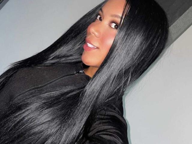 DaimondJohnson with long black shiny hair, smiling, wearing a black zip-up top, showing a cute face with brown eyes and pink lipstick, in a selfie.