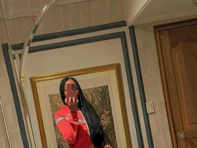 DaimondJohnson, com longas tranças pretas, camisa vermelha, segurando o celular em um selfie no espelho, mostrando o bumbum no reflexo, em pé em um quarto com uma porta de madeira e um quadro..