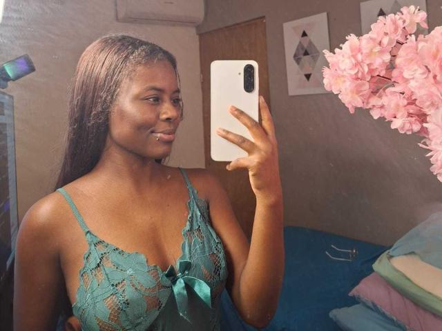 Kairaa stands in a room, taking a mirror selfie with a white smartphone. She has dark brown skin and long, straight black hair. She wears a teal lace camisole with a bow at the center. Her facial expression is neutral, with a small silver barbell piercing in her lower lip. Pink cherry blossom flowers are in the foreground. The reflection shows her upper body. The image is slightly blurred.