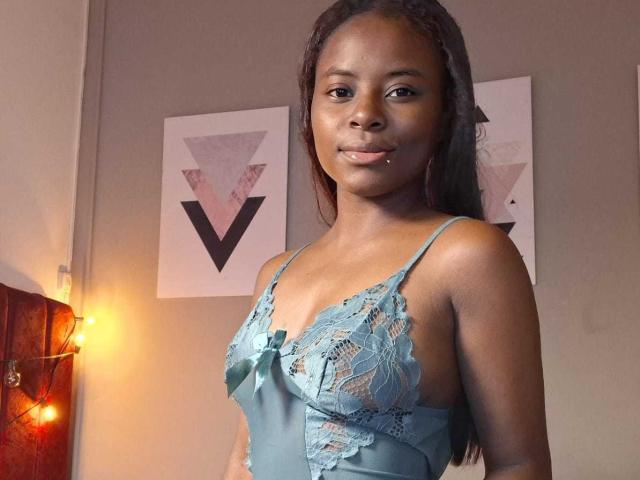 Kairaa stands in a room, wearing a light blue lace lingerie top with a bow. Her dark brown skin contrasts with the delicate lace pattern. She has long, straightened hair and a small piercing on her right lower lip. Two geometric art prints with pink, black, and white triangles are on the gray wall behind her. A string of warm yellow lights is on the left. The room is softly lit, highlighting her confident, relaxed expression.