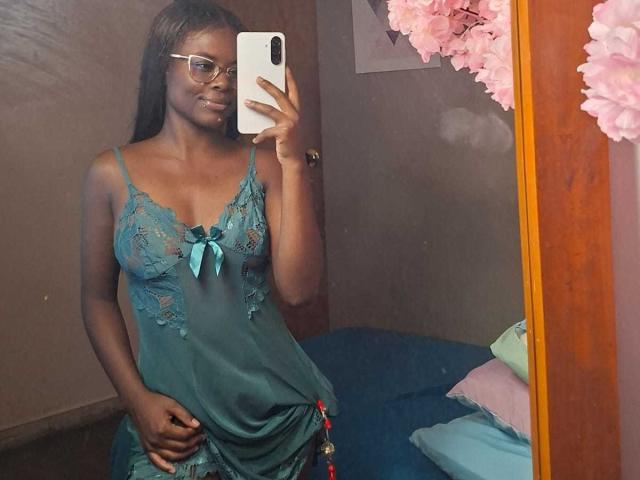 Kairaa with dark skin, glasses, long brown hair, wearing a teal lace babydoll with a blue ribbon, holding a phone in a mirror selfie, showing her small tits.
