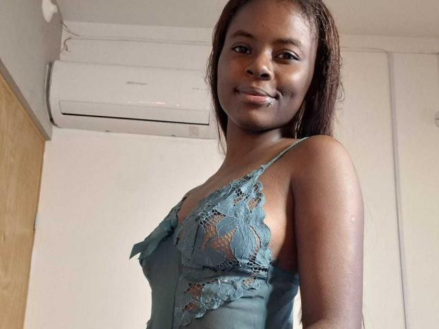 Kairaa with dark skin, wearing a teal lace lingerie top, showing her small tits, smiling slightly, standing in a room with an air conditioner above her.