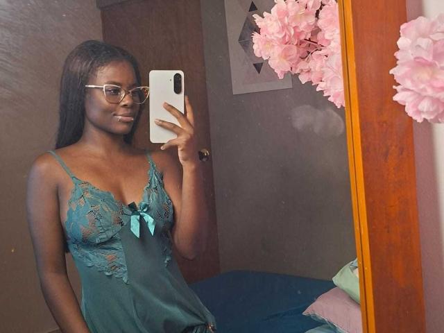 Kairaa with dark skin, long black hair, glasses, and blue lace lingerie, showing her tits in a mirror selfie. Pink flowers in the mirror. Her phone is white.