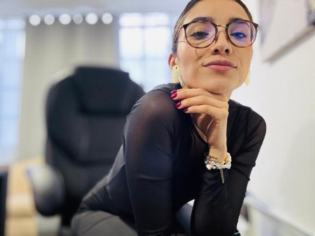 AdharaCooperr, a woman with light brown skin and dark hair in a bun, wears glasses with purple-tinted lenses and a black long-sleeve shirt. She rests her chin on her hand, smiling slightly. She has red-painted nails and a bracelet with a cross on her wrist. Her earrings are gold and small. The background is blurred with a black office chair and light from a window. The image has a soft focus and a slight lens flare.