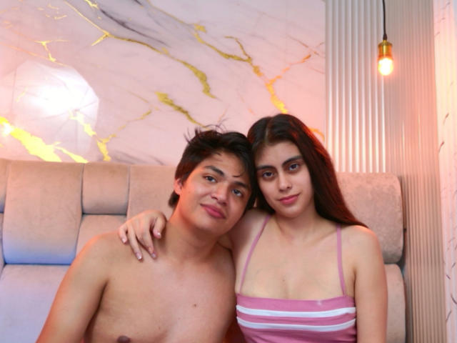 SofiNdLogan with short brown hair, shirtless, and SofiNdLogan with long brown hair, pink striped top, sitting on beige couch, arm around each other, looking at camera, soft smile.