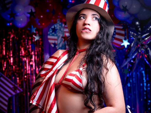 NicolSky with long black wavy hair, wearing a red and white striped bikini top and hat, looking up with a slight smile, her tits visible, in a festive, colorful setting.