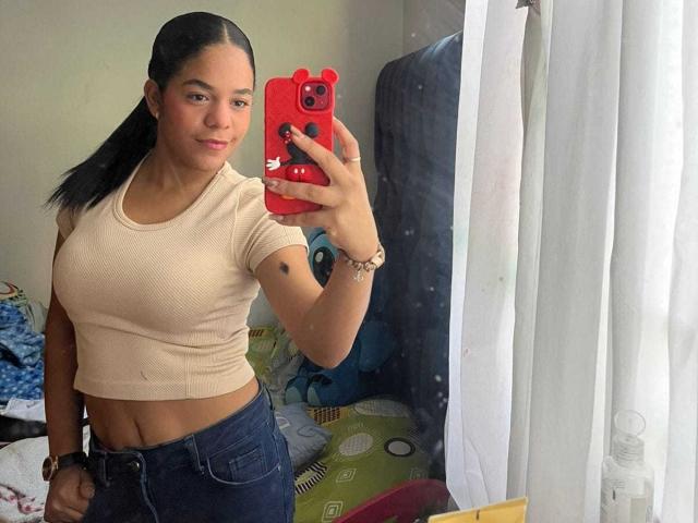 TiffanyyJoness with black hair in a ponytail, wearing a beige crop top and blue jeans, holding a red phone, taking a mirror selfie, showing her tits and midriff.