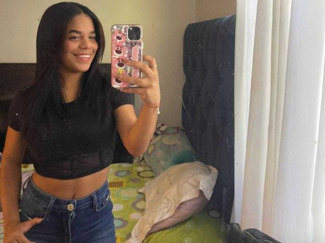 TiffanyyJoness with long black hair, smiling, wearing a black crop top and blue jeans, holding a pink phone, standing in a bedroom, showing a mirror selfie.