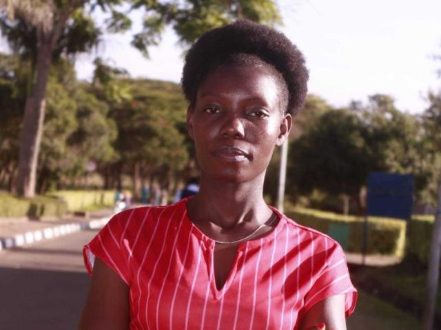 JulianaMirabel with dark skin, short afro, wearing a red striped shirt, looking straight at the camera, sunlight on her face, street in the background.