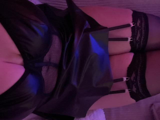 Claraax69 in black lingerie with garter straps, sheer mesh, and lace, showing her tits and upper thighs, in a purple-lit bedroom.