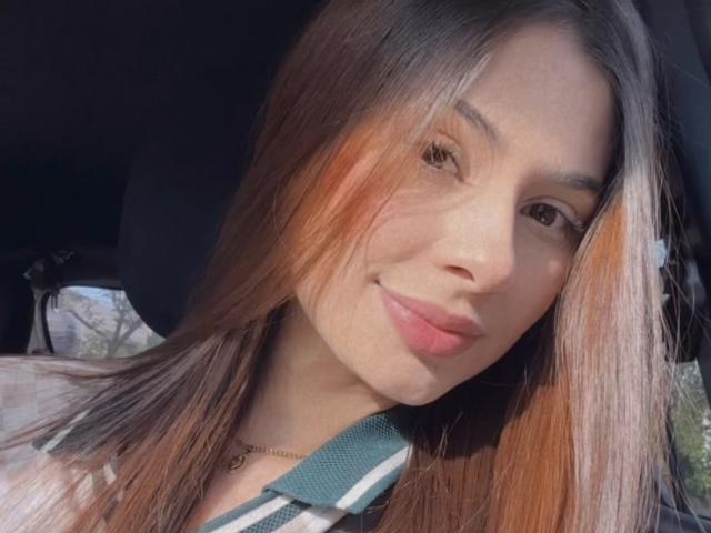 NikoleFoster has long brown hair with orange highlights, a slight smile, and is wearing a green and white striped top with a seatbelt across her chest.
