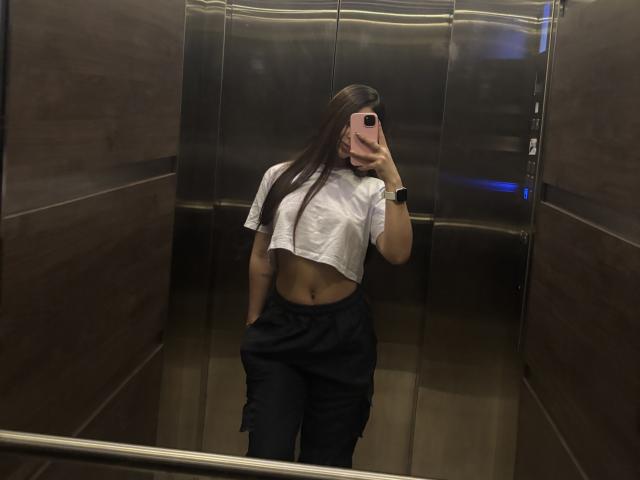NikoleFoster with long brown hair, wearing a white cropped shirt, black pants, and a black wristwatch, taking a mirror selfie in a elevator. Her hand's in her pants.