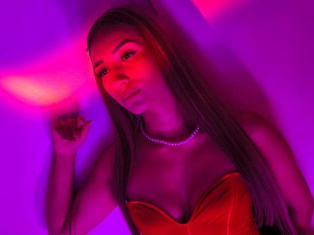 TanniaMoon with long blonde hair, red velvet strapless top, and pearl necklace, glowing under purple and pink lights, looking slightly to the side with a subtle smile.