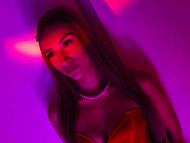 TanniaMoon with long brown hair, red dress, pearl necklace, and pink-purple lighting, looking at camera with subtle smile, soft shadow on right side.