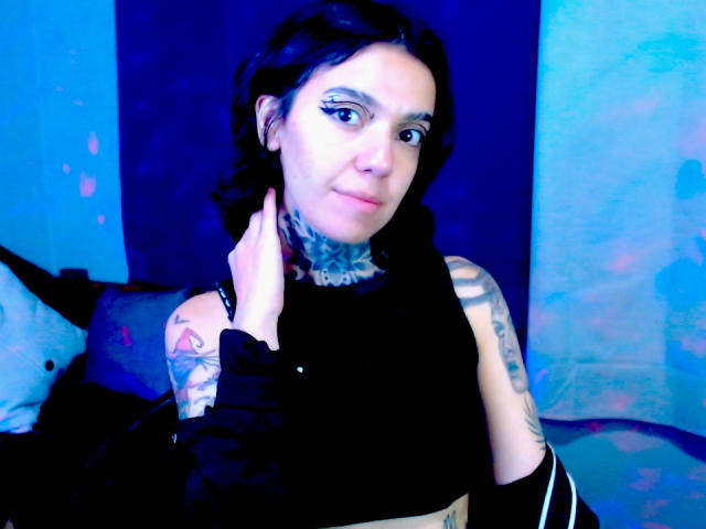 LannieGonzales with dark hair, tattoos on neck and arm, black sleeveless top, blue lighting, hand on neck, slight smile, blue background, visible eyeliner, subtle makeup.