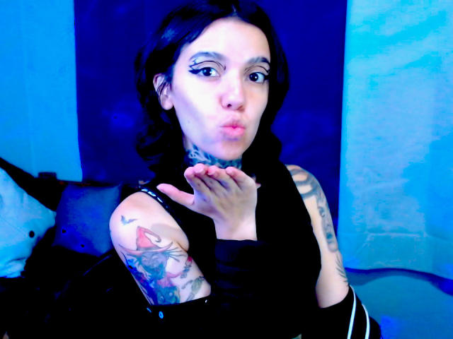 LannieGonzales with dark hair, pouty lips, and tattoos, wearing a black sleeveless top, hand near chin, blue lighting, visible tattoos on arm and neck.