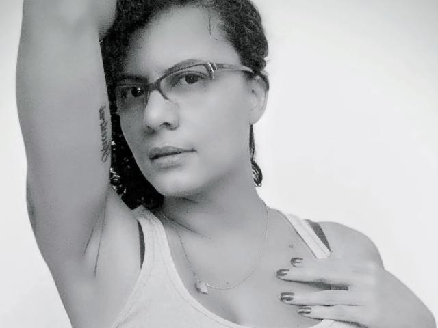 RosannaBlue with glasses, raised arm, tank top, visible tattoo on arm, hand on chest, nails painted dark, medium skin, curly hair, necklace, black-and-white photo.