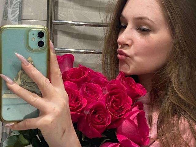 PamelaXu with pouty lips, long brown hair, pink roses, holding a green phone with a beige case, wearing pink, with long, pointed nails, looking at herself.