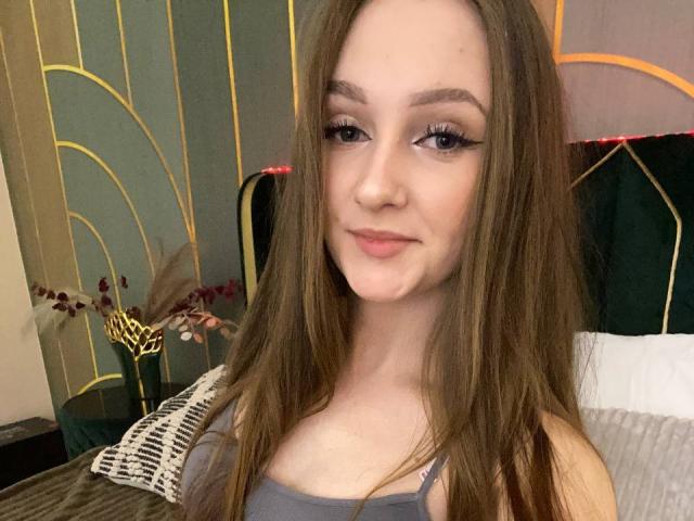 PamelaXu has long brown hair, eyeliner, and a subtle smile. She's wearing a gray tank top, showing her small tits, with a wicker pillow in the background.