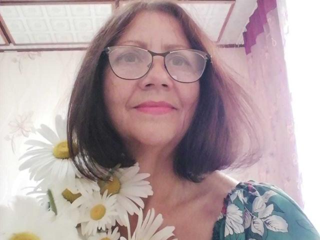 OlgaHottie with glasses, medium brown bob, pink lipstick, holding white daisies with yellow centers, wearing a floral green top, smiling slightly.