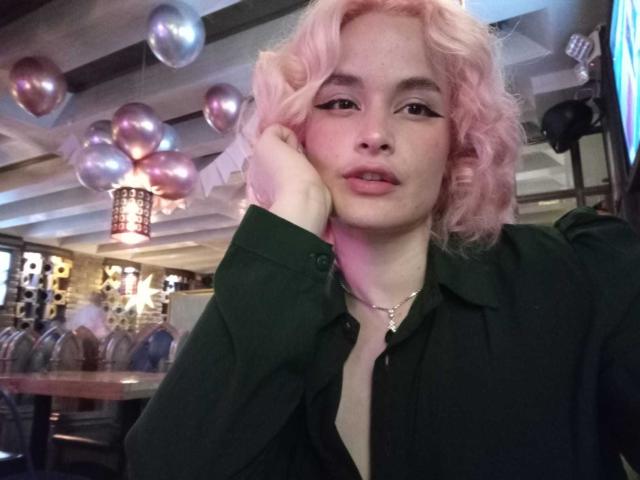 DanaStone with pink curly hair, green shirt, black chest, hand on cheek, slightly open mouth, silver balloon reflections, casual, confident, soft pink lighting, necklace, relaxed expression.