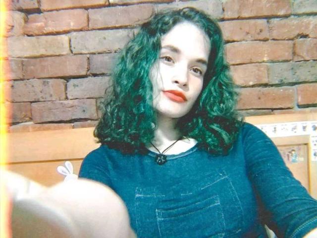 DanaStone has curly green hair, red lipstick, and wears a dark blue denim shirt with pockets. She's looking at the camera with a slight smile.