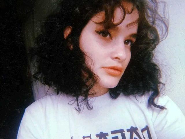 DanaStone with curly black hair, pale skin, and winged eyeliner, wearing a white shirt, has a serious expression and slightly parted orange lips. Her ass is not visible.