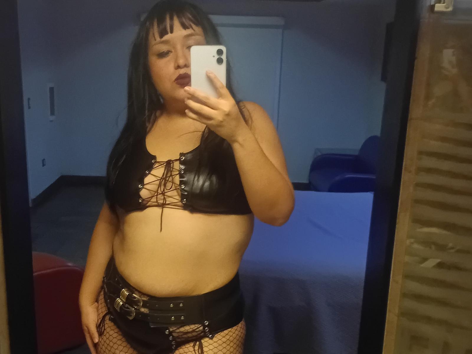 Ixtab with black leather crop top, lace-up front, showing her tits, black leather shorts, belt with buckles, fishnet stockings, and dark lipstick, taking a mirror selfie.