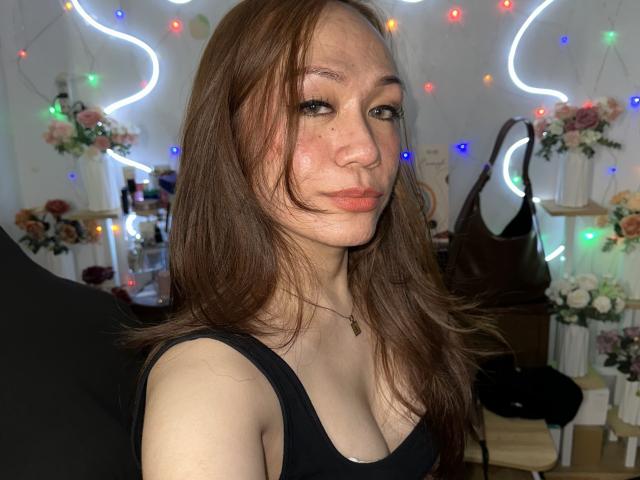 TsJade with brown hair, fair skin, and small tits, wearing a black top, has a pouty expression, and is in a room with colorful lights and flowers.