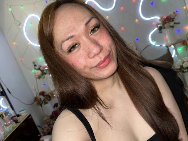 TsJade with long brown hair, light skin, and small tits, smiling slightly, wearing a black strap top, neon lights in the background, and colorful fairy lights on the wall.