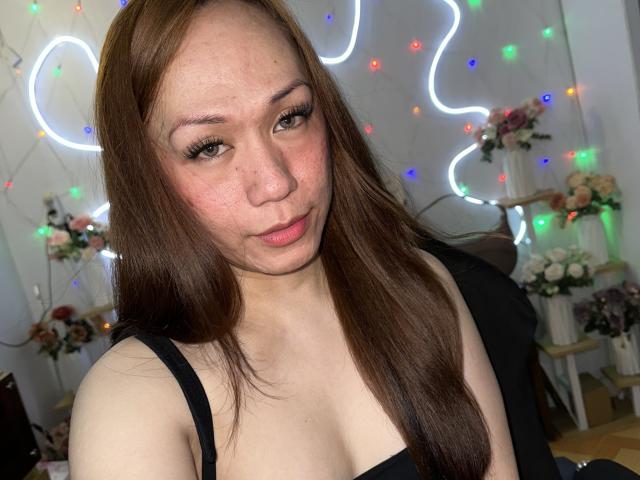 TsJade has brown hair, light skin, and is wearing a black top. She has small tits and is making a slightly pouty face. Neon lights and flowers are in the background.