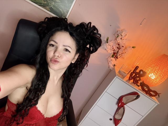 MollyBrunett with curly black hair, red lace lingerie, puffy lips, sitting in black chair, chest visible, white dresser with red heels and orange lamp in background.