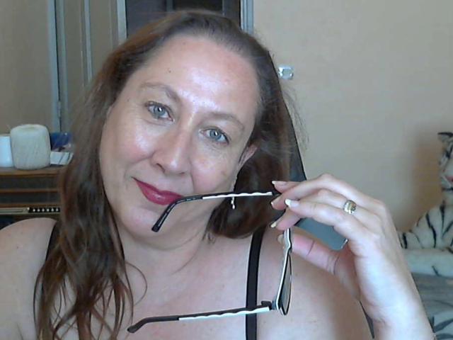 XMyriamX, middle-aged woman with brown hair, holding glasses in her mouth, smiling with red lipstick, wearing black strap top, showing her tits, ring on right hand, in bedroom.
