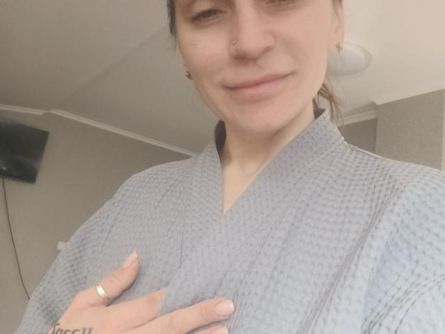 AliaAnita with a light gray waffle robe, slight smile, nose piercing, ring on left hand, hand on chest, brown hair in a ponytail, subtle makeup, soft lighting, indoor.