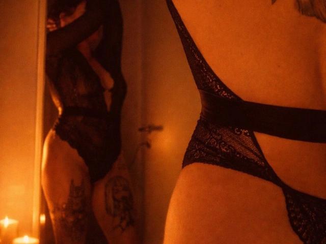 HeyLina-hot in black lace lingerie, showing a tattoo on her thigh and a side view of her butt, with a mirror reflection showing her back and arm.