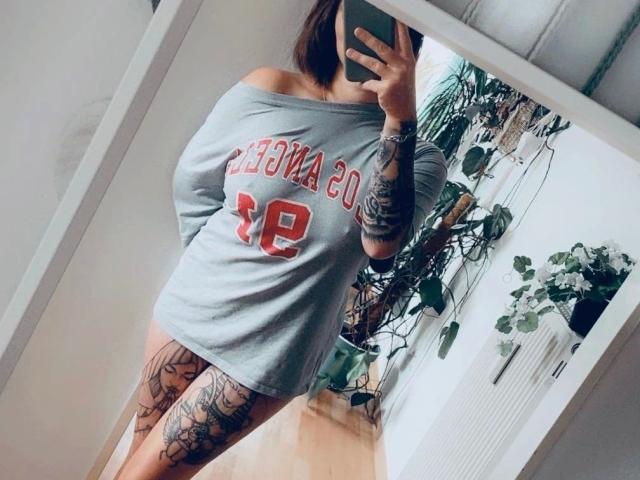 HeyLina-hot with off-shoulder grey "Chico's" shirt, showing tattoos on arms and legs, holding phone in mirror selfie, wood floor, green plants in background.