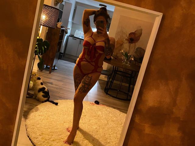 HeyLina-hot in red strappy lingerie, showing off her tits and ass, with tattoos on her thigh, taking a mirror selfie in a cozy room.
