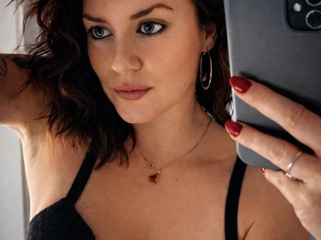 HeyLina-hot with wavy brown hair, blue eyes, and red nails, wearing black lingerie, holding a phone, silver hoop earrings, and a necklace with a red pendant.