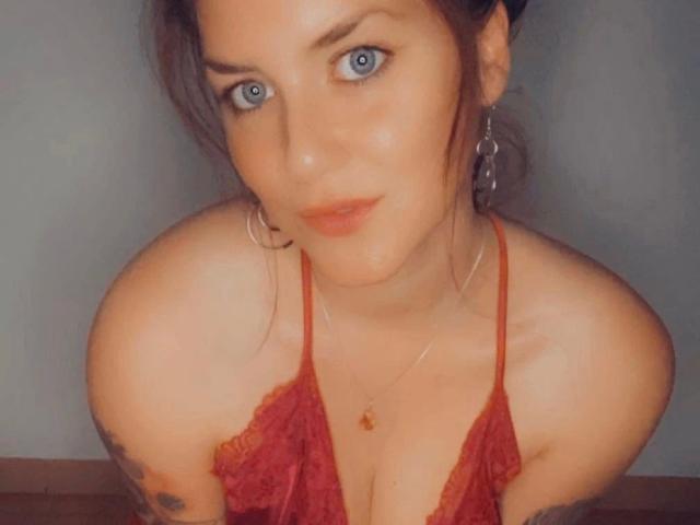 HeyLina-hot with blue eyes, wearing a red lace halter top, showing her tits, has tattoos on her arms, hoop earrings, and a necklace with a small pendant.