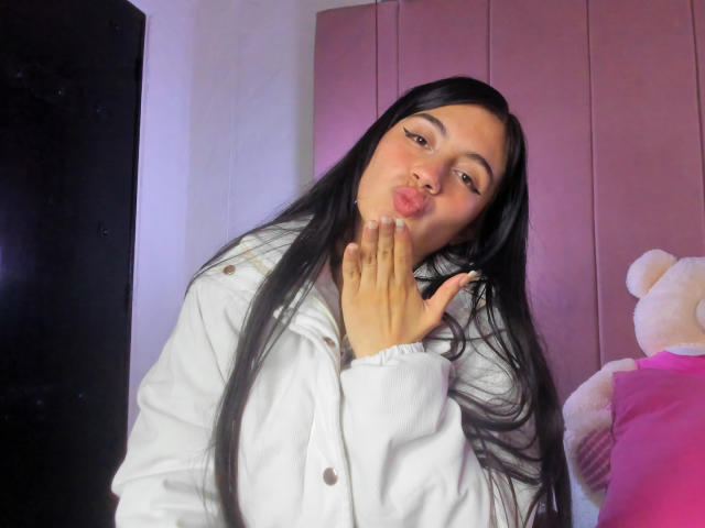 YinYAngluar with long black hair, puffy white jacket, kissing her fingers, soft pink background, fluffy pink teddy bear on the right. Her hand is near her slightly open mouth.