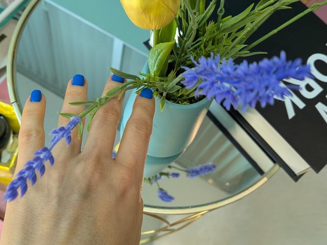 AuroraStar's hand with bright blue nail polish touches a blue flower in a light blue vase, yellow tulip, and green foliage on a glass table.