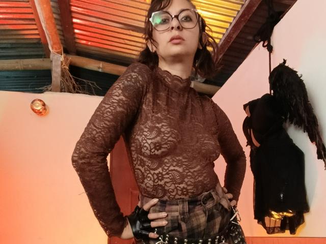 SerenaXYZ with glasses, wearing a sheer brown lace top, her small tits visible, hands on hips, black plaid skirt, dark hair, looking down, in a dimly lit room.
