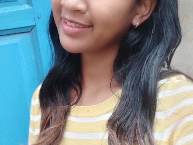 Close-up of LoderFist, a young woman with medium brown skin and wavy black hair with subtle blonde highlights. She wears a yellow and white striped shirt and small hoop earrings. Her lips are slightly parted in a gentle smile, showing white teeth. The blue door in the background contrasts with her warm skin tone and yellow shirt. The image focuses on her face and upper chest.