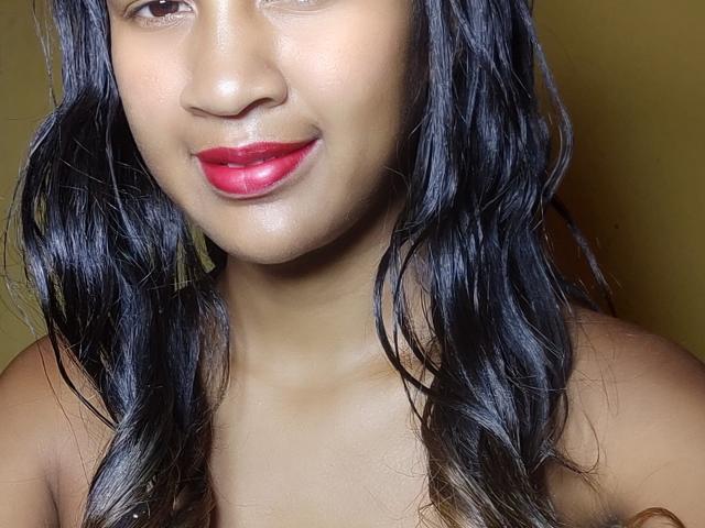LoderFist has dark, wavy hair, brown skin, and red lipstick. She's smiling with her eyes slightly closed, and she's topless, showing her shoulders.