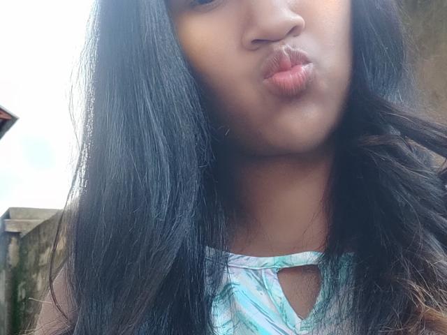 Close-up of LoderFist, a woman with long, straight black hair, pouting her lips. She has medium brown skin and is wearing a light blue and white patterned top with a small keyhole cutout. The texture of her hair is smooth and shiny. The image is slightly overexposed, giving a bright, almost washed-out look to her face. The background is out of focus, with a hint of a building and a roof on the left.